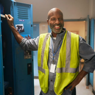 Man standing in locker room with a blue locker open and leaning on the door with a yellow safety vest on smiling at the camera.